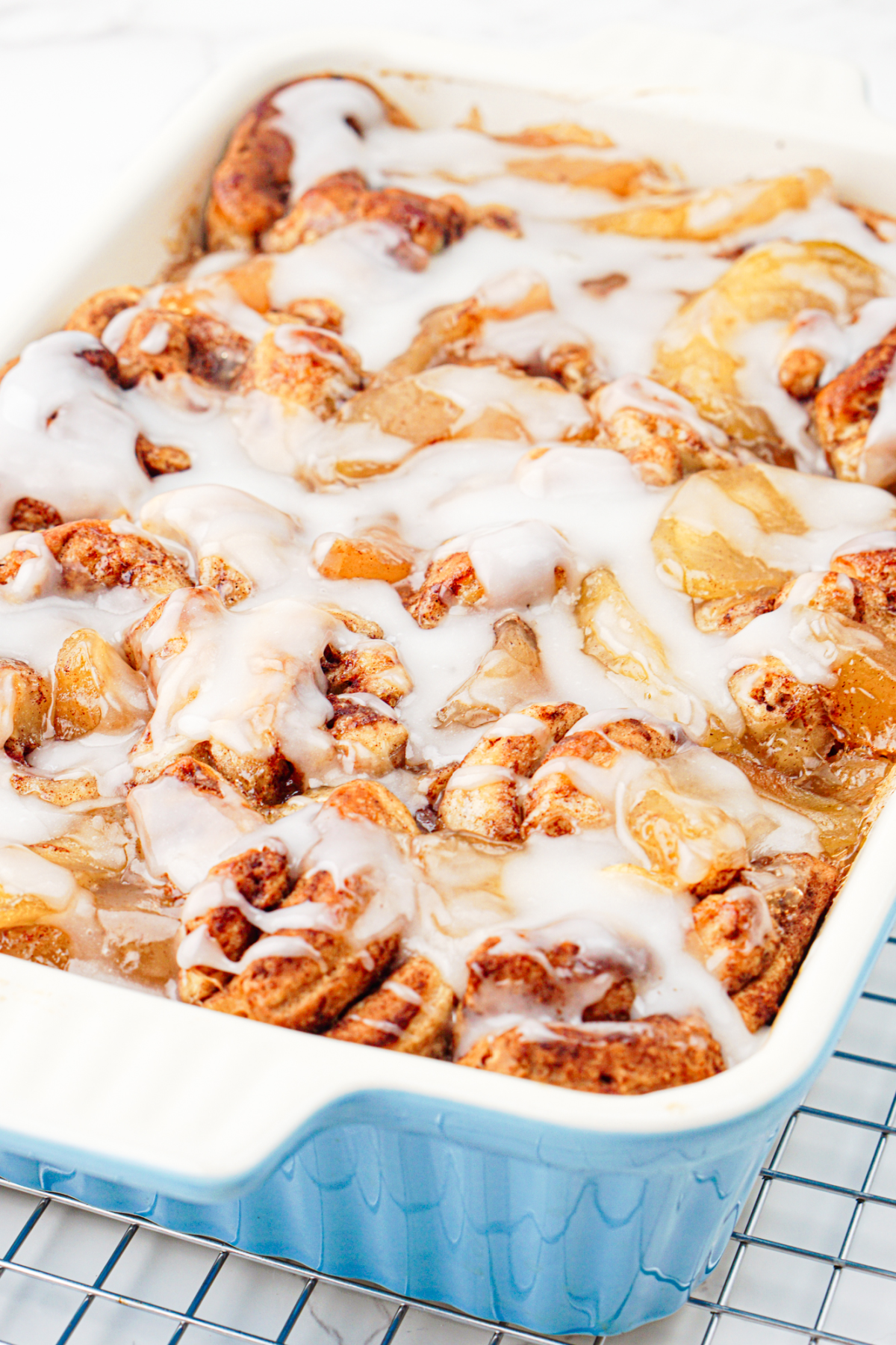 A blue baking dish filled with apple cinnamon rolls topped with white icing, fresh out of the oven and resting on a cooling rack.