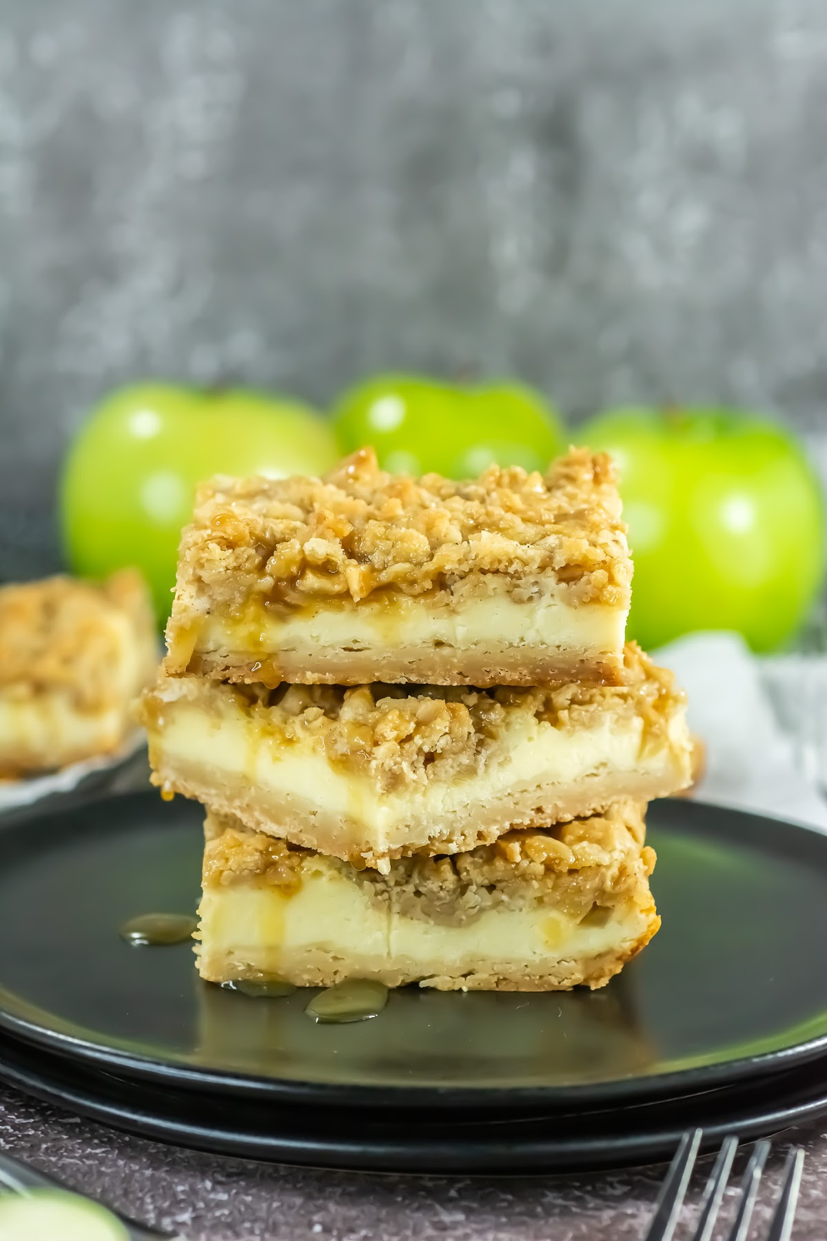 Two caramel apple cheesecake bars are stacked on a black plate, with green apples and more bars visible in the blurred background.