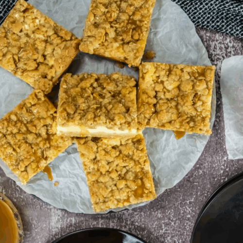 Seven caramel apple cheesecake bars with oat crumble are arranged on a parchment-lined plate, accompanied by caramel sauce and a green apple visible in the top right corner.