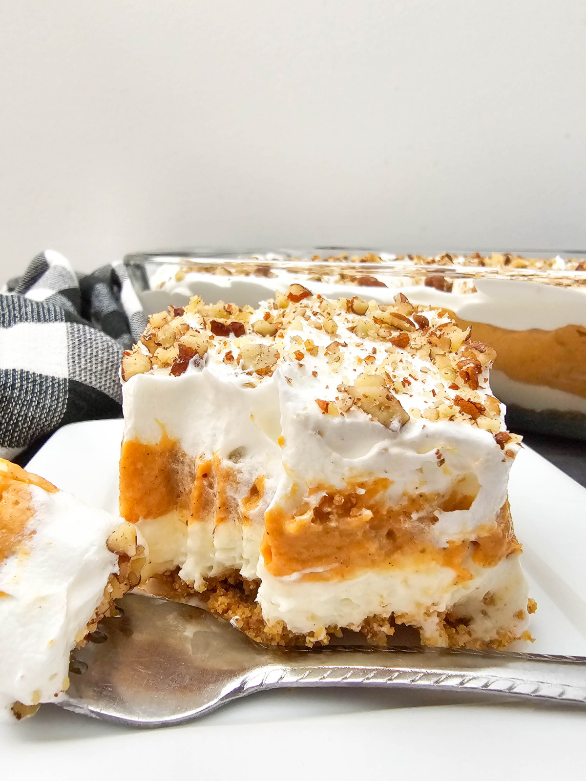 A slice of pumpkin lush dessert with a crumbly crust, creamy filling, pumpkin layer, whipped topping, and chopped nuts on a white plate with a spoon.