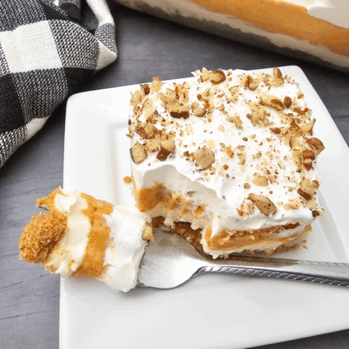 A square of pumpkin lush dessert with whipped topping and chopped nuts sits on a white plate; a fork holds a bite beside it. A checkered cloth is nearby.
