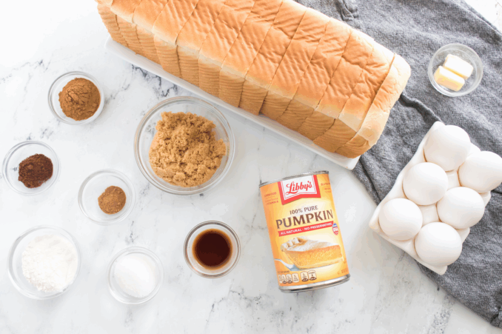 Sliced white bread, eggs, canned pumpkin, brown sugar, butter, vanilla, and various spices arranged on a white surface—perfect for making a delicious pumpkin French toast bake.