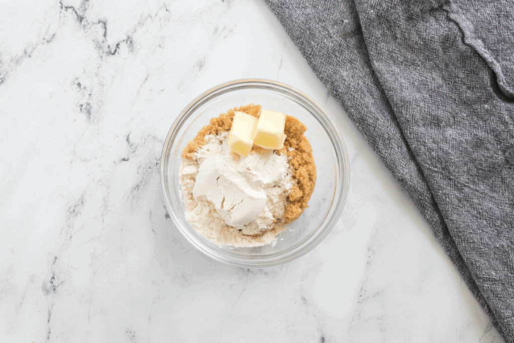 A glass bowl with brown sugar, flour, and butter cubes—ingredients perfect for a pumpkin French toast bake—sits on a marble surface next to a gray cloth.