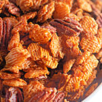 Close-up of a bowl filled with a crunchy snack mix containing pecans and cereal pieces coated in a caramelized glaze.