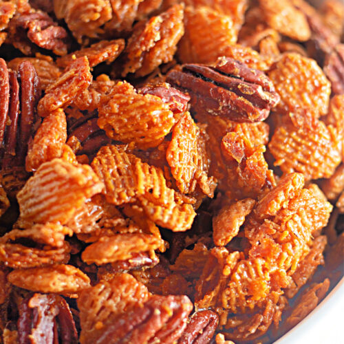 Close-up of a bowl filled with a crunchy snack mix containing pecans and cereal pieces coated in a caramelized glaze.