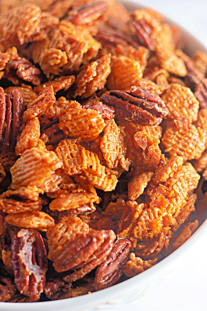 Close-up of a bowl filled with a crunchy snack mix containing pecans and cereal pieces coated in a caramelized glaze.