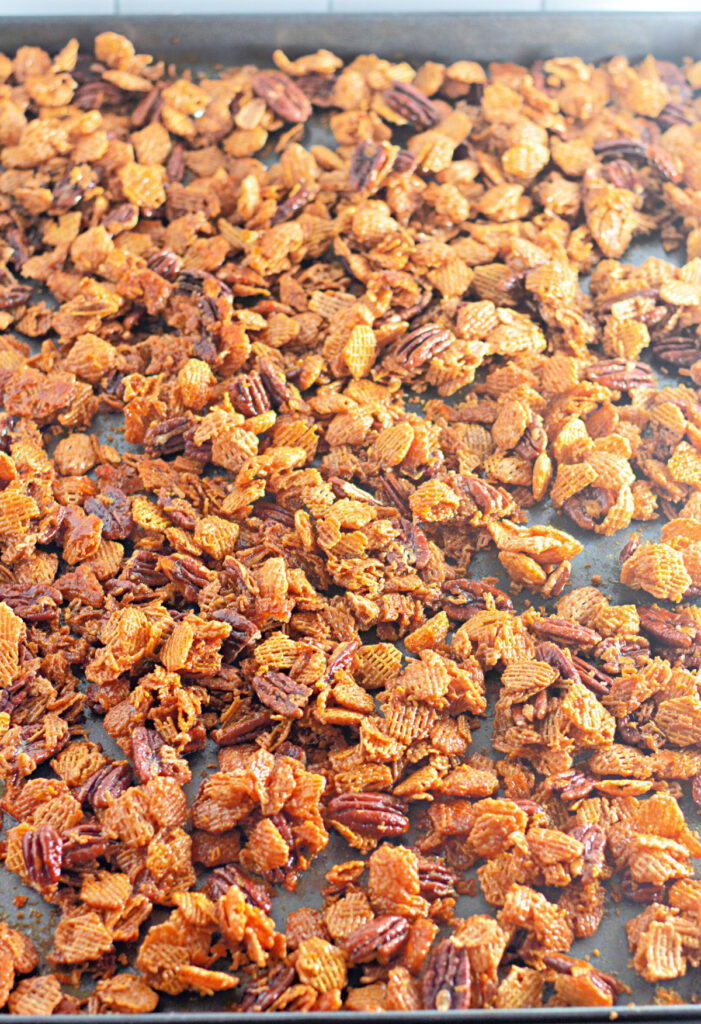 A baking tray filled with a golden-brown snack mix, including cereal pieces and pecans, evenly spread out and toasted.