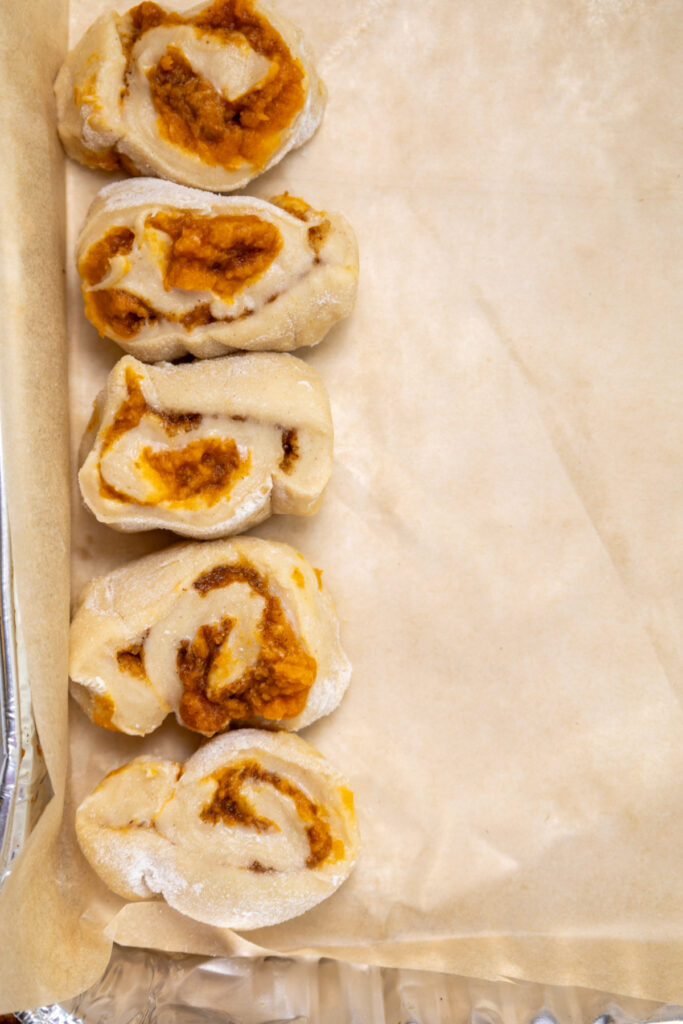 Four unbaked pumpkin cinnamon rolls with beautiful swirls are arranged in a vertical line on a parchment-lined baking tray.