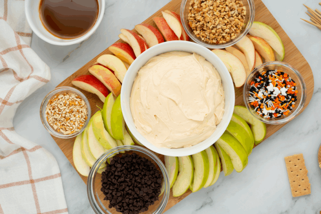 A wooden board with sliced red and green apples, a bowl of creamy caramel apple dip in the center, and small bowls of chocolate chips, chopped nuts, and sprinkles arranged around it.