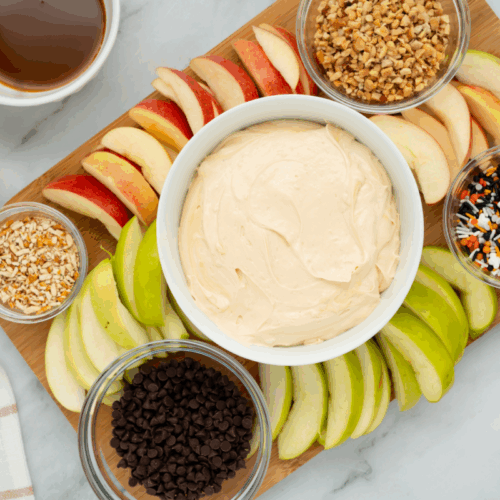 A wooden board with sliced red and green apples, a bowl of creamy caramel apple dip in the center, and small bowls of chocolate chips, chopped nuts, and sprinkles arranged around it.