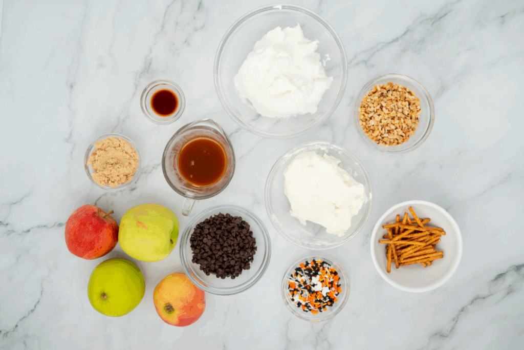 Various ingredients in bowls, including apples, chocolate chips, chopped nuts, brown sugar, cream cheese, whipped topping, caramel sauce for caramel apple dip, pretzel sticks, and Halloween sprinkles arranged on a marble surface.