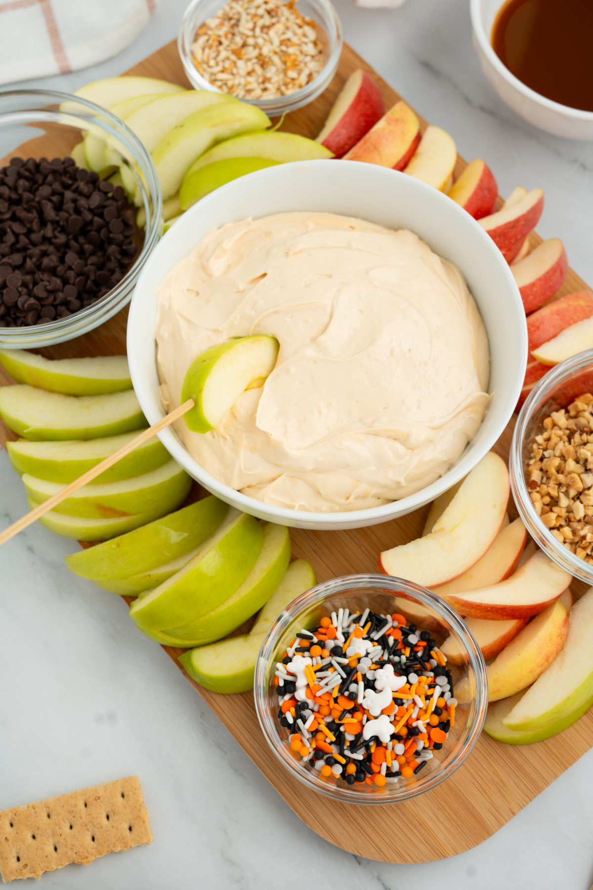 A platter with sliced green and red apples surrounds a bowl of caramel apple dip in the center, with small bowls of toppings like chocolate chips, nuts, and sprinkles.