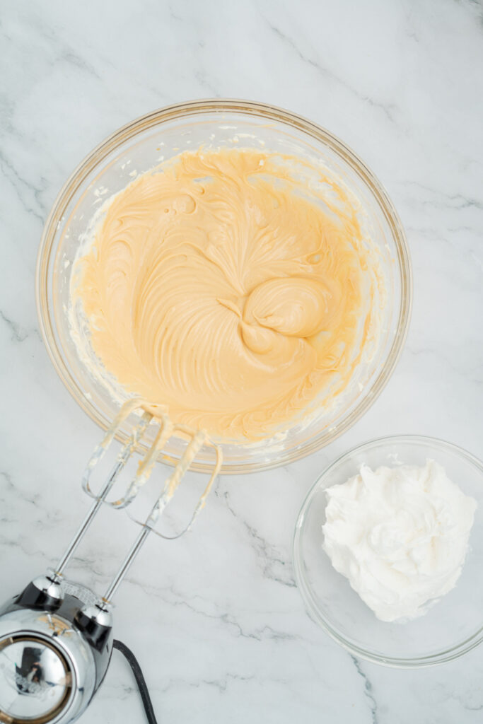 A glass bowl with creamy caramel apple dip batter and a hand mixer beside a smaller bowl containing whipped cream, all on a marble surface.
