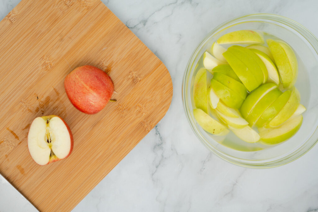 A red apple is cut in half on a wooden cutting board, while sliced green apples soak in a bowl of water on a marble surface, ready to pair with caramel apple dip.