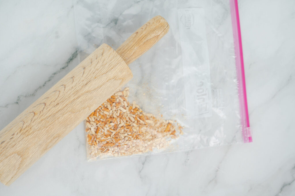 A wooden rolling pin crushing dried onions inside a clear plastic zip-top bag on a marble surface, perfect for adding crunch to your caramel apple dip.