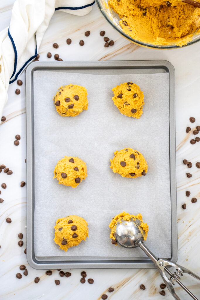 A baking tray lined with parchment paper holds six scoops of cookie dough with chocolate chips. A metal scoop, bowl of dough, and scattered chocolate chips are nearby.