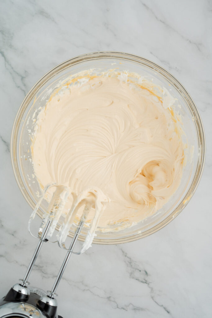 A glass bowl of light tan whipped caramel apple dip frosting is shown on a marble surface with a hand mixer resting inside.