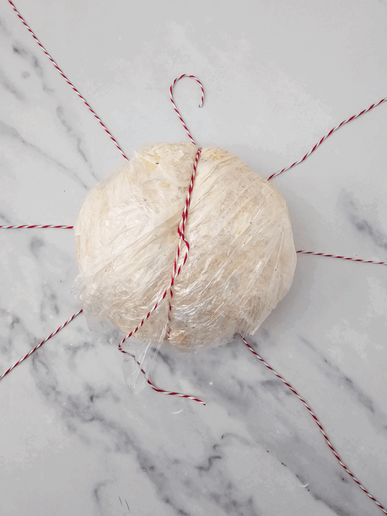 A pumpkin cheese ball wrapped in plastic wrap with red and white string tied around it rests on a marble surface.
