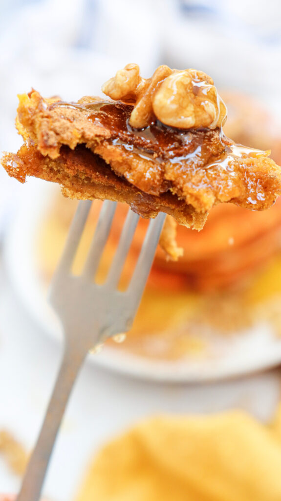 A close-up of a fork holding a bite of pancakes topped with walnuts and drizzled with syrup.