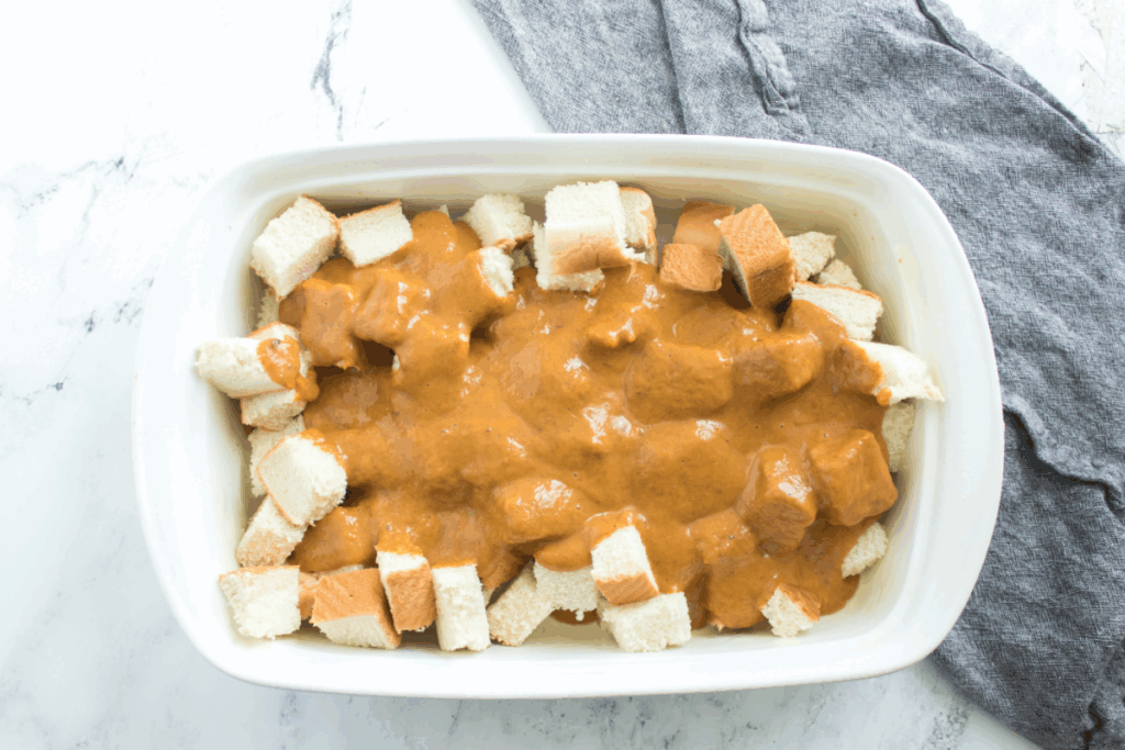A rectangular white baking dish with cubed white bread topped with a thick brown sauce, resembling a pumpkin french toast bake, sits on a white marble surface next to a gray cloth.