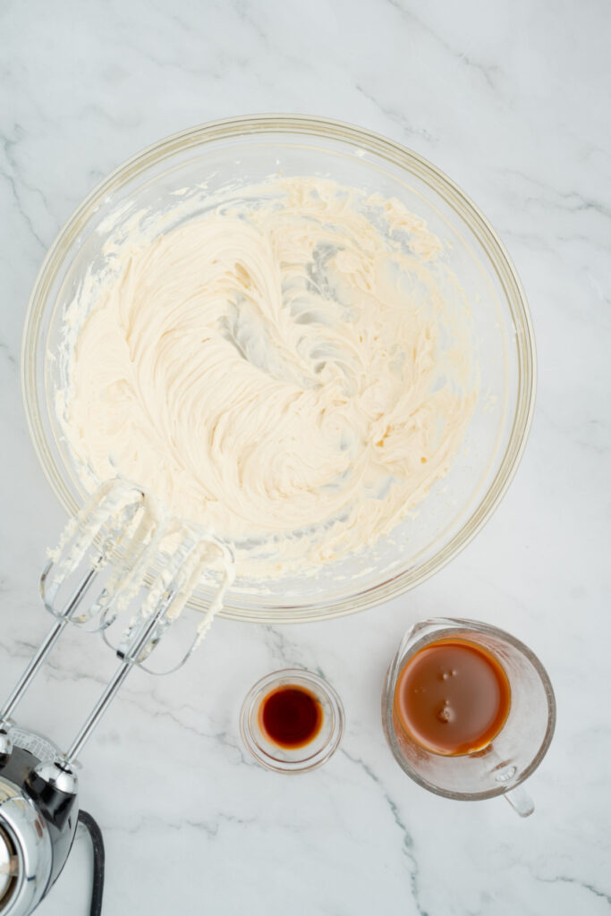 A glass bowl of whipped cream mixture with an electric hand mixer, next to a small glass of vanilla extract, a measuring cup of coffee, and caramel apple dip on a marble countertop.