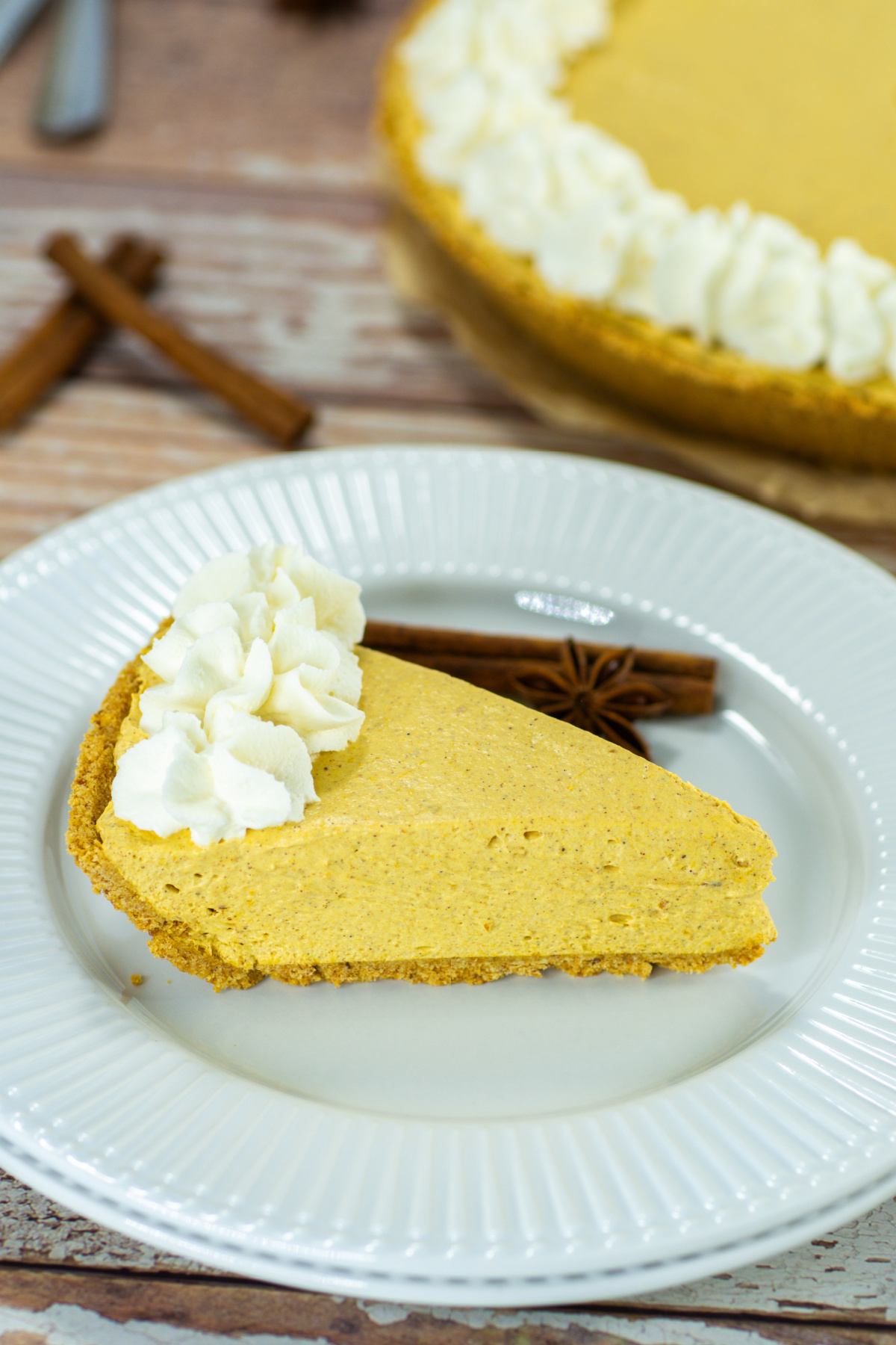 A slice of pumpkin pie with whipped cream on a white plate, garnished with cinnamon sticks, and the rest of the pie in the background.