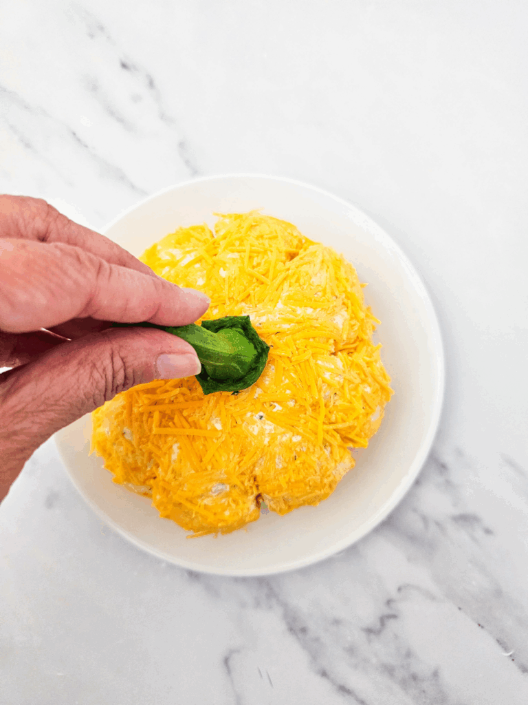 A hand places a green leaf on top of a pumpkin cheese ball covered in shredded cheddar on a white plate, set on a marble surface.