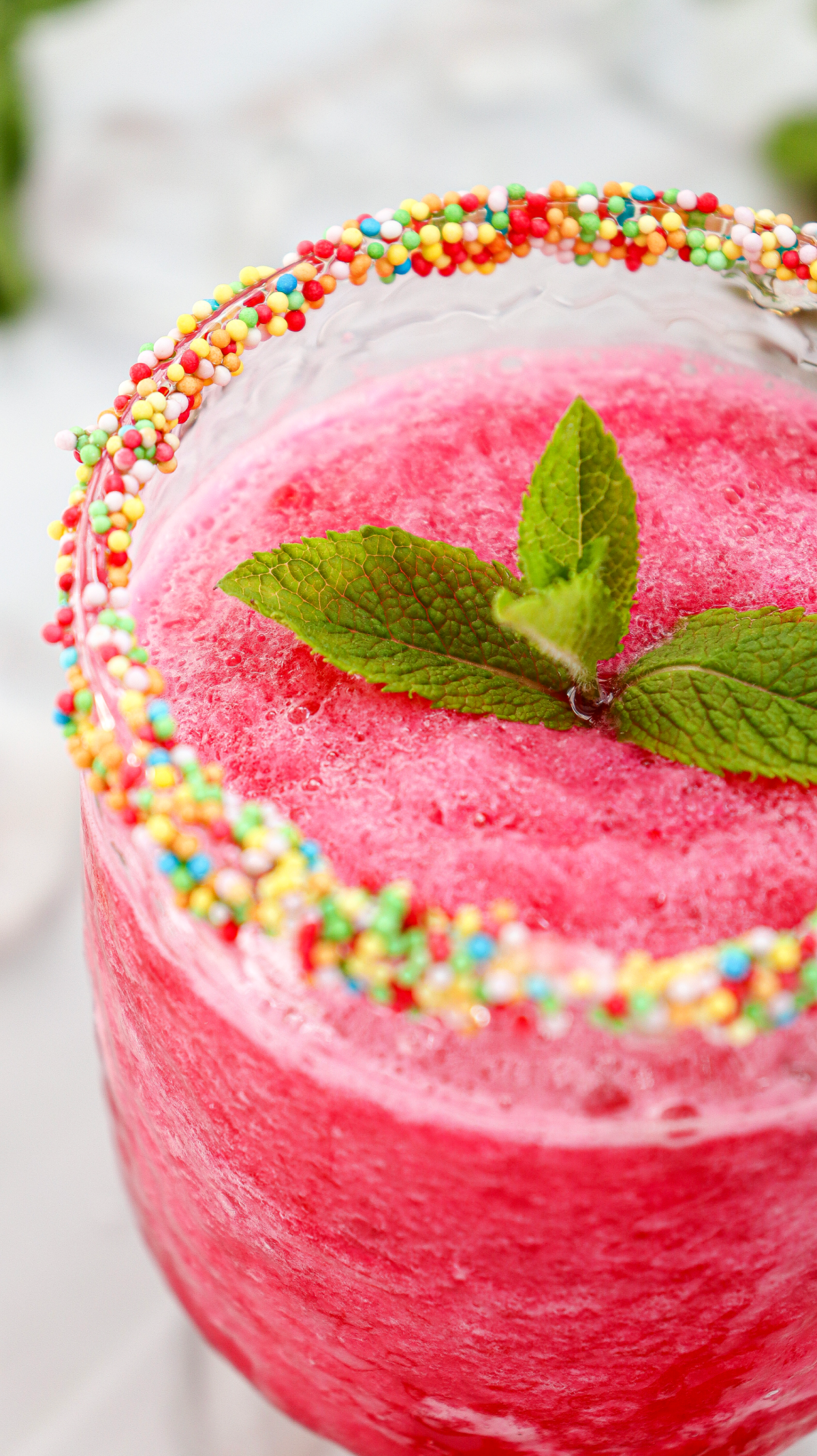 A close-up of a vibrant watermelon slushie topped with mint leaves, with the glass rim decorated in colorful sprinkles.