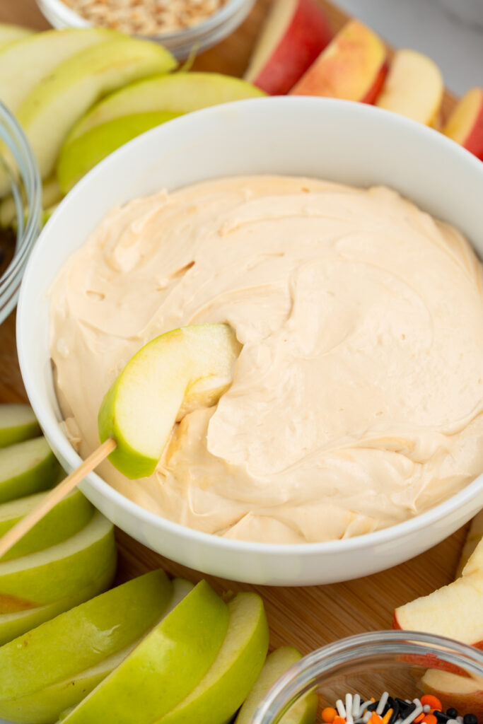 A white bowl filled with creamy caramel apple dip is surrounded by sliced green and red apples; a green apple slice is dipped into the bowl.