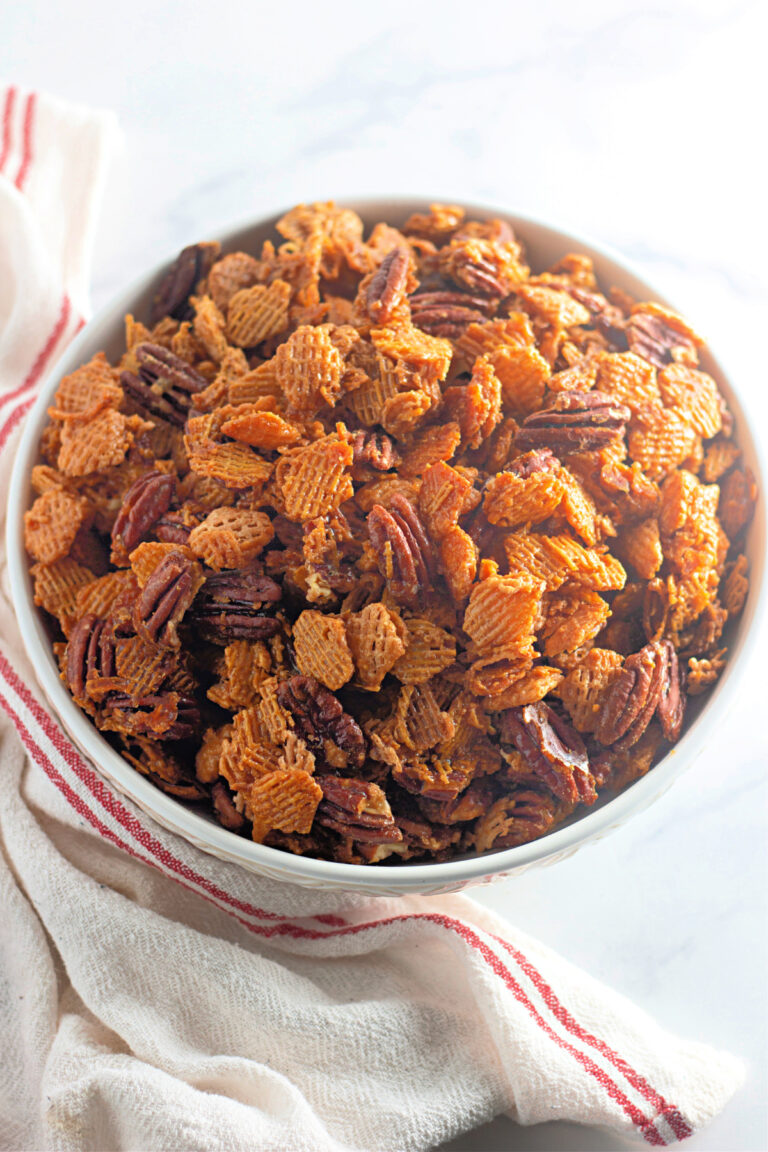 A white bowl filled with a mixture of Chex cereal and pecans sits on a white cloth with red stripes, set on a light surface.