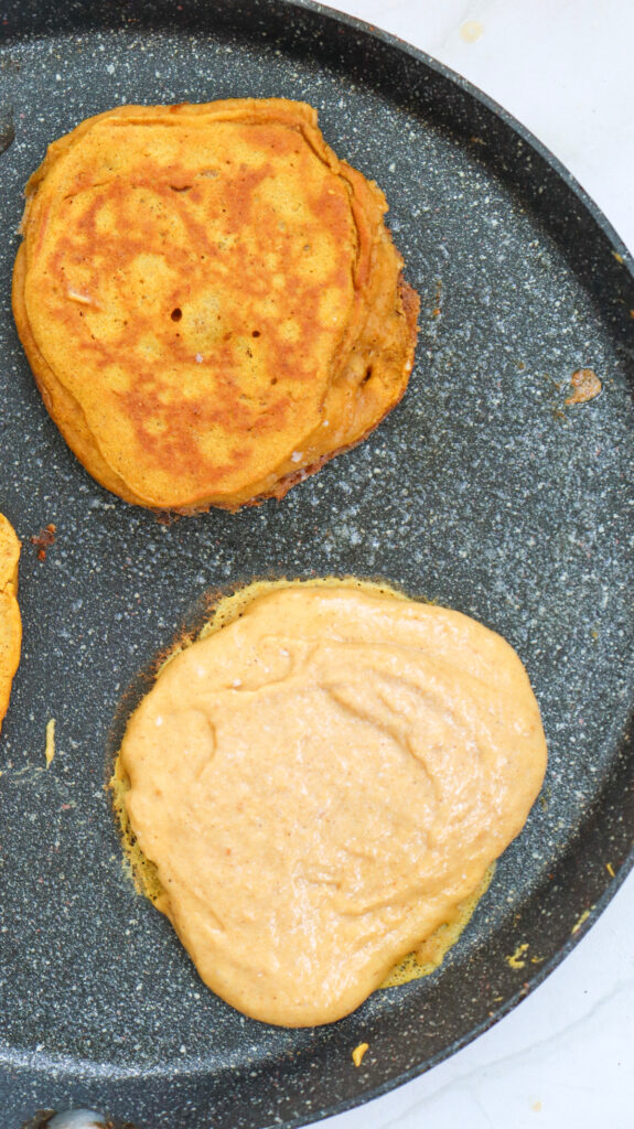 Two pancakes are cooking in a nonstick pan; one is golden brown and cooked, while the other is still raw batter.