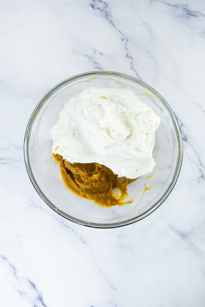 A glass bowl containing whipped cream and pumpkin puree on a white marble surface.