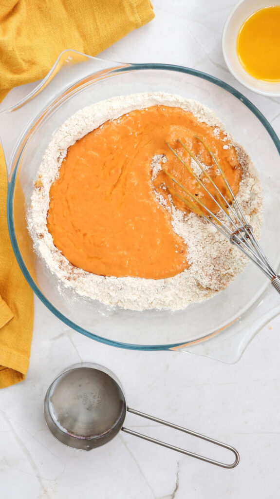 A glass bowl with flour and orange batter being mixed with a whisk, next to a measuring cup and a bowl of melted butter on a white countertop.