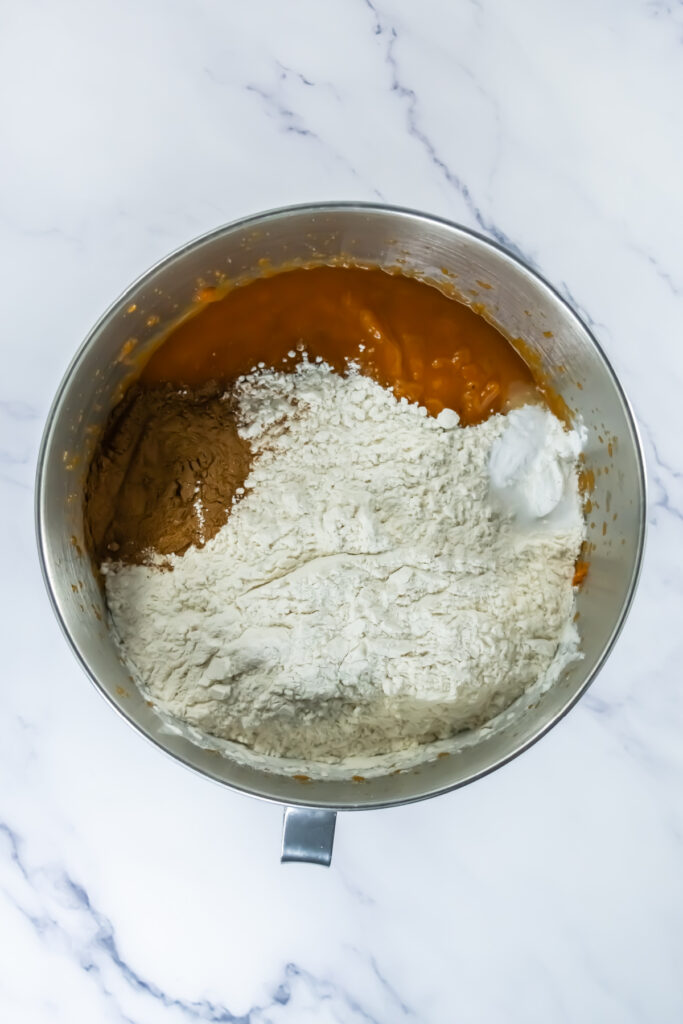 A mixing bowl containing pumpkin puree, flour, cocoa powder, and baking powder on a white marble surface.
