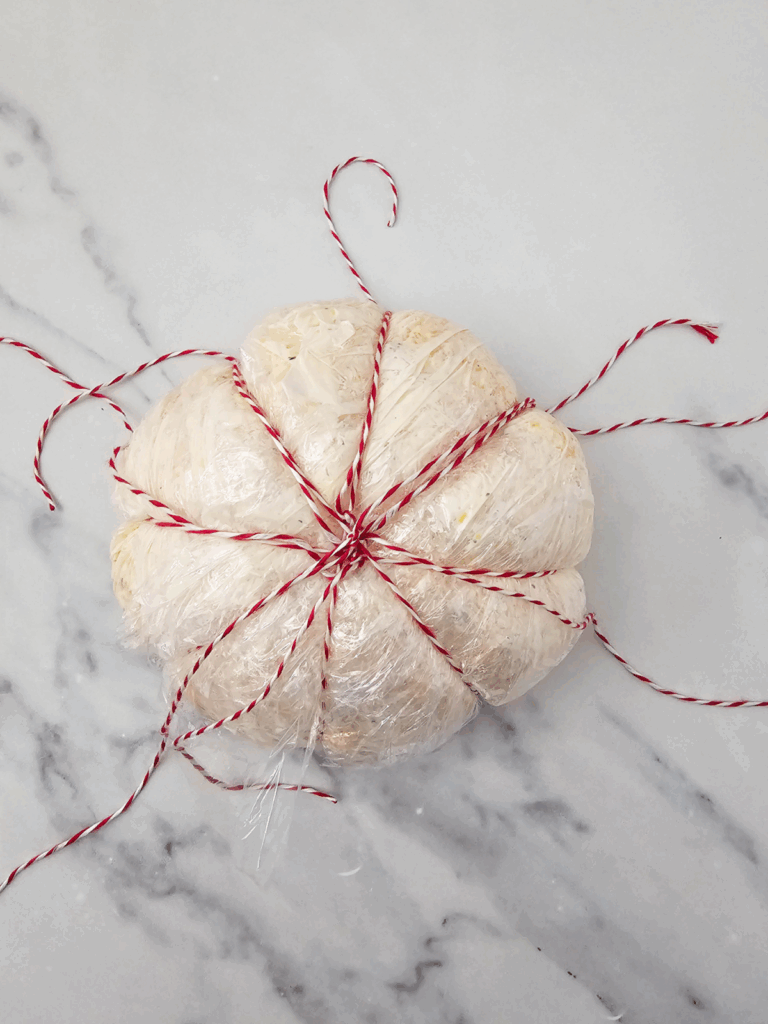 A pumpkin cheese ball, wrapped in plastic wrap and tied with red and white string, sits on a white marble surface.