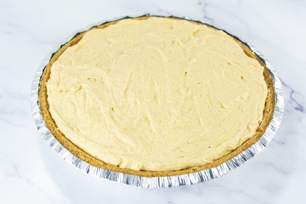 A whole, uncut pie with a light tan filling sits in a foil pie tin on a white marble surface.