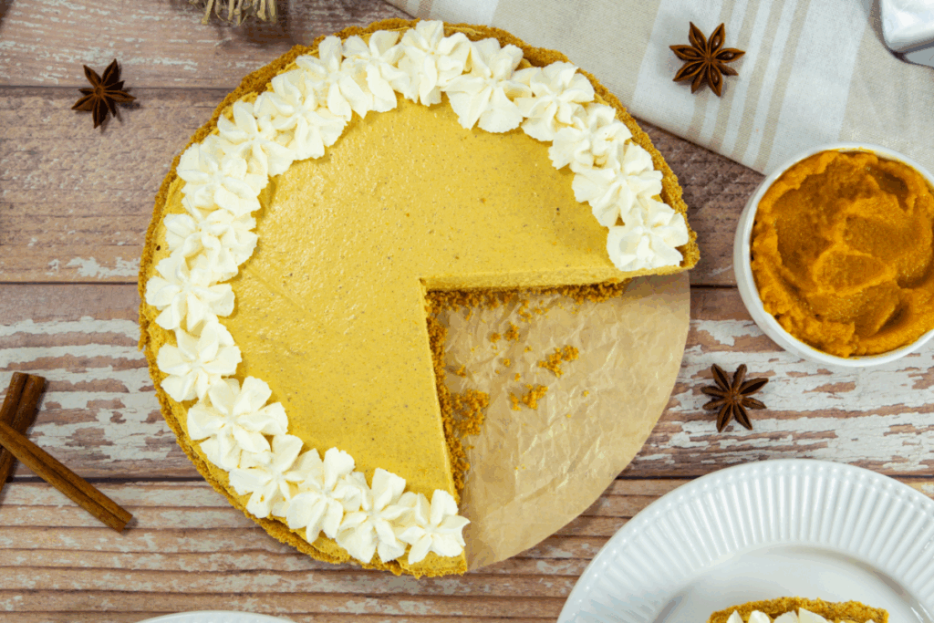 A pumpkin pie with a missing slice, topped with whipped cream rosettes on half of the crust, sits on parchment paper next to a small bowl of pumpkin puree and star anise.