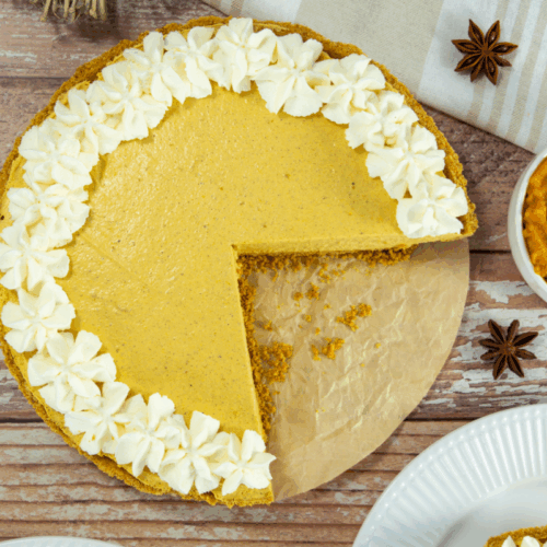 A pumpkin pie with a missing slice, topped with whipped cream rosettes on half of the crust, sits on parchment paper next to a small bowl of pumpkin puree and star anise.