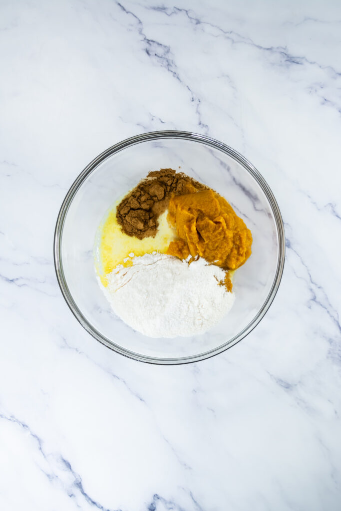 A glass bowl containing flour, brown sugar, melted butter, and pumpkin puree on a white marble surface.
