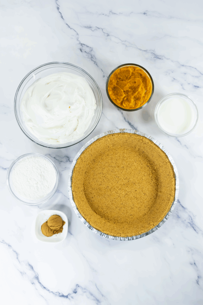 A graham cracker pie crust, whipped topping, pumpkin puree, powdered sugar, milk, and spices arranged on a white marble surface.