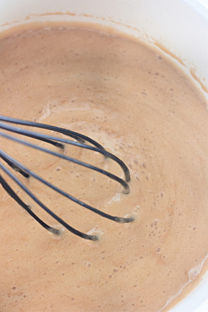 A metal whisk stirring a bowl of light brown batter or mixture.