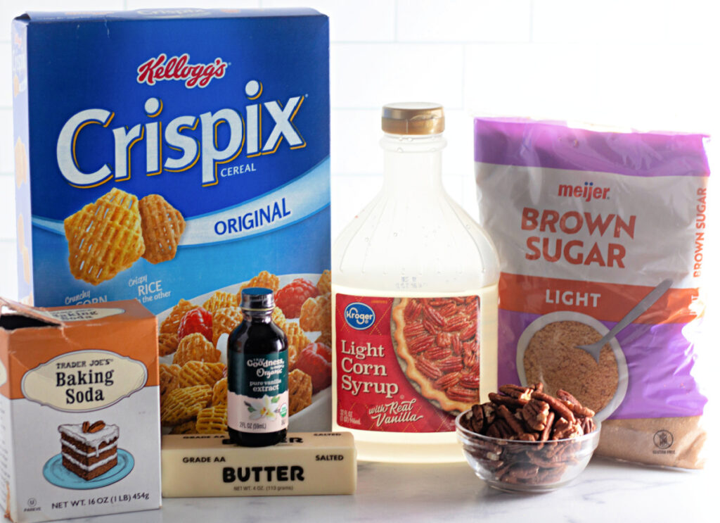 A box of Crispix cereal, brown sugar, light corn syrup, pecans, vanilla extract, butter, and baking soda arranged on a white surface.