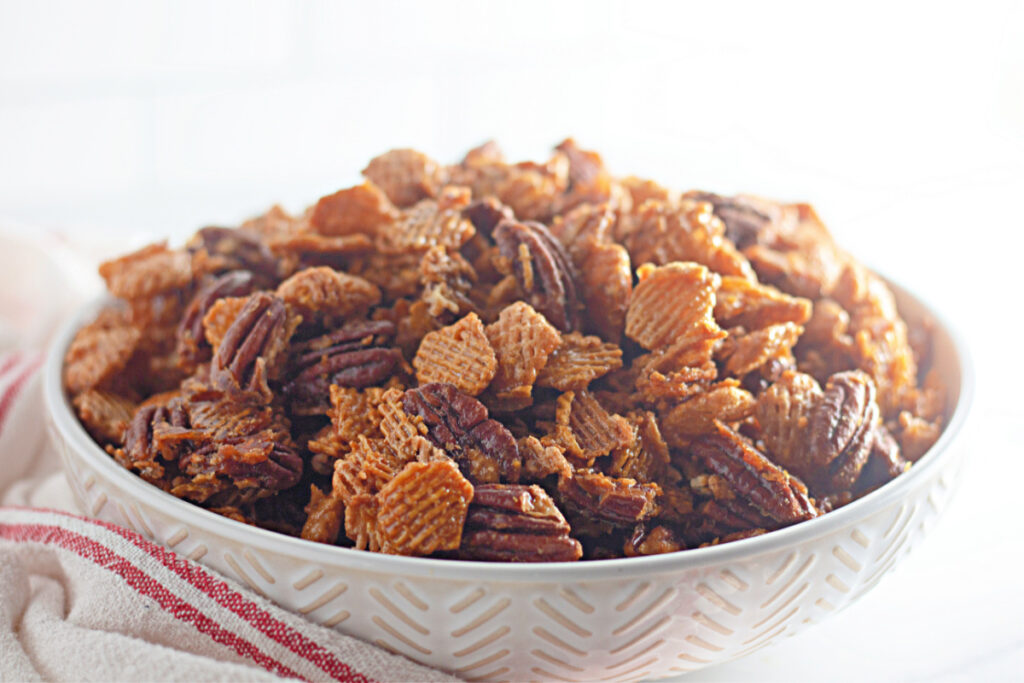 A white bowl filled with a mix of cereal squares and pecans sits on a white surface with a red-striped towel beside it.