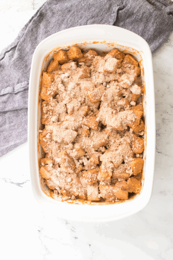 A white baking dish filled with cubed pieces of bread coated in a caramel-colored sauce and topped with a crumbly streusel, this pumpkin French toast bake sits on a marble surface next to a gray cloth.