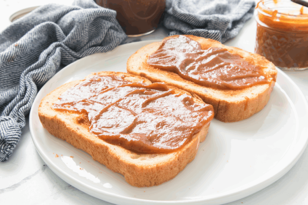 Two slices of bread with a thick layer of pumpkin butter sit on a white plate, next to a striped cloth and jars of spread.