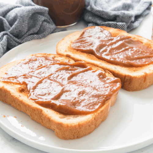 Two slices of bread with a thick layer of pumpkin butter sit on a white plate, next to a striped cloth and jars of spread.