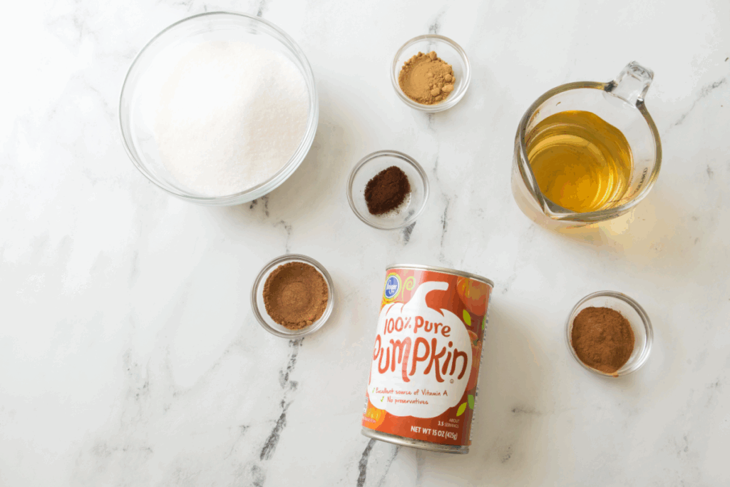 Ingredients on a marble surface: a can of pure pumpkin, a bowl of sugar, spices in small bowls, and a measuring cup with a light brown liquid—perfect for making homemade pumpkin butter.
