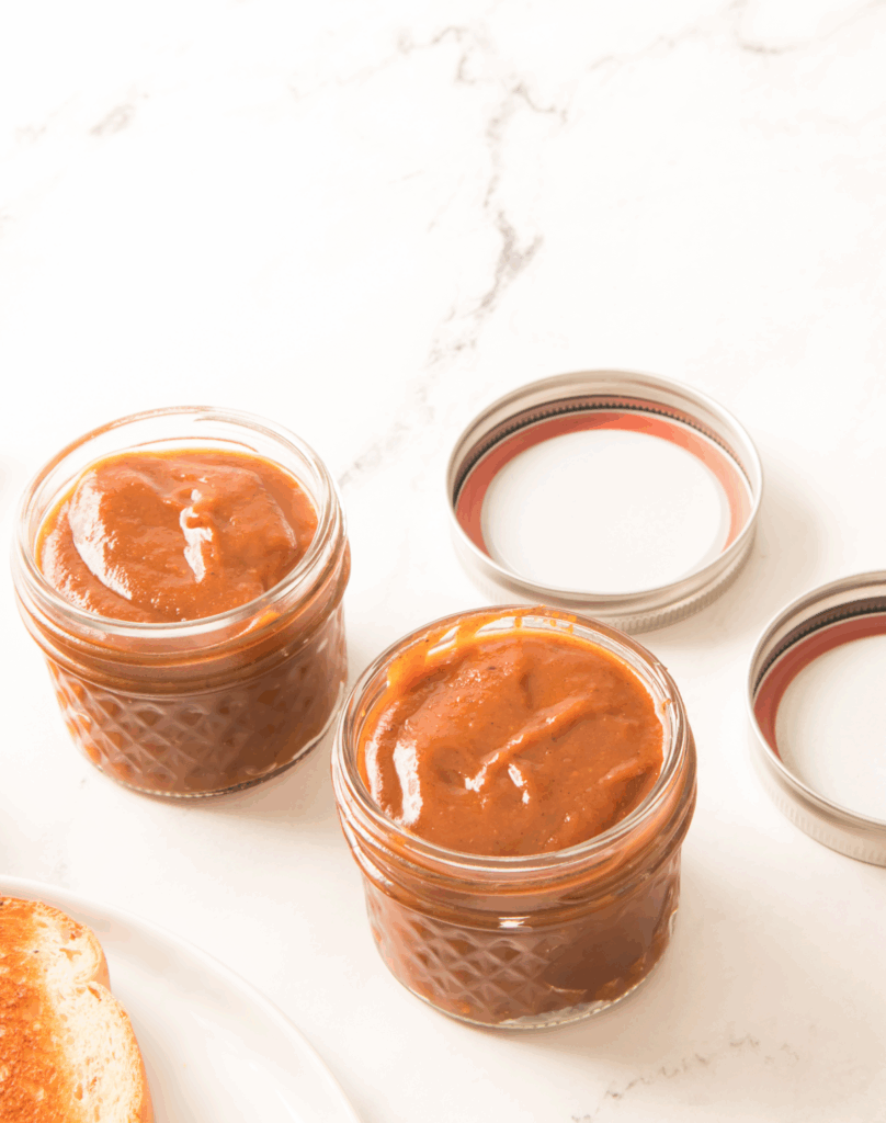 Two small glass jars filled with thick pumpkin butter sit on a white marble surface beside two jar lids and part of a plate with toast.