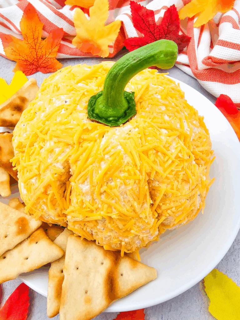 A pumpkin cheese ball shaped like a pumpkin, coated in shredded cheese and finished with a green stem, sits on a white plate surrounded by crackers and autumn leaves for the perfect fall appetizer.