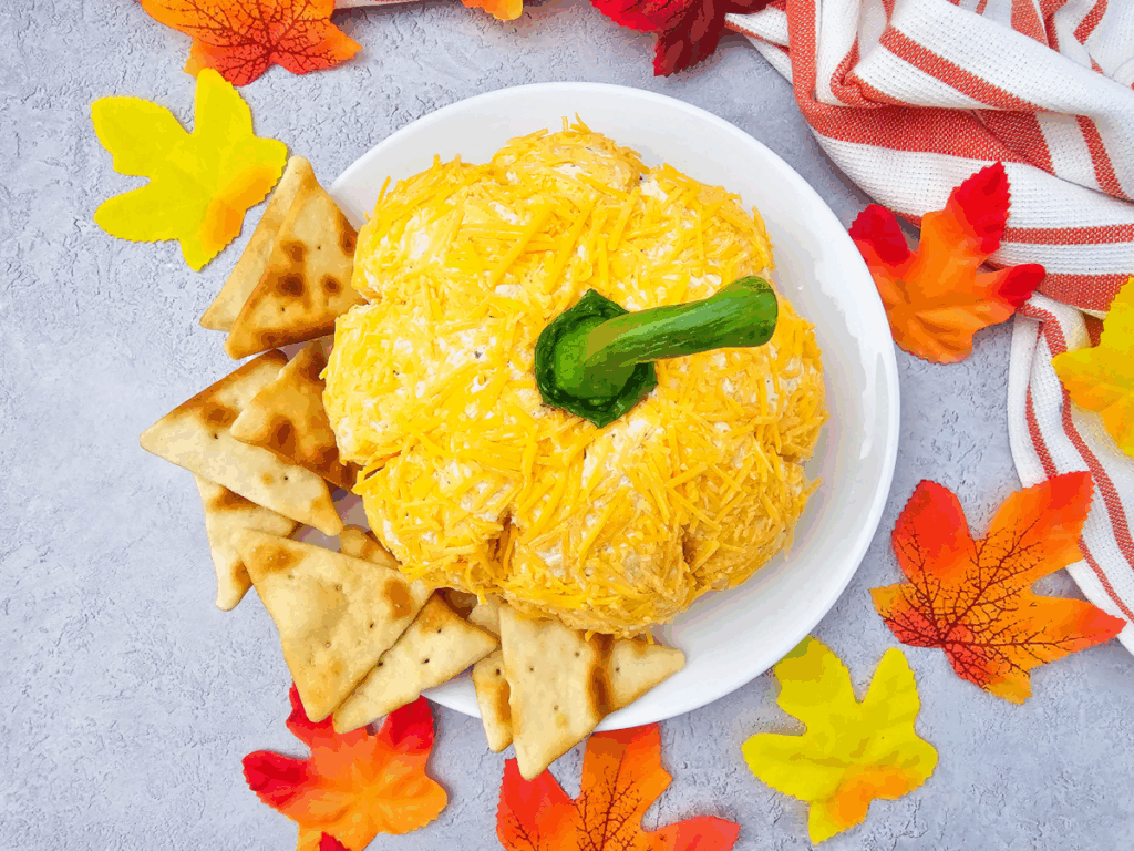A pumpkin cheese ball with a green stem is served on a white plate with pita chips, surrounded by colorful artificial fall leaves and a striped towel.